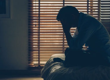 foto de uma homem em quarto escuro sentado à beira da cama, com as mãos na cabeça e olhando para baixo