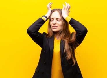 Foto de uma mulher em fundo amarelo com as duas mãos na cabeça demonstrando ansiedade.