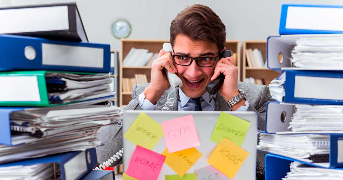 foto de homem no trabalho atendendo dois telefones e com pilhas de relatórios ao lado. seu semblante é de desespero