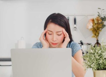 Foto de uma mulher em frente ao computador com as mãos na cabeça e semblante estressado e cansado