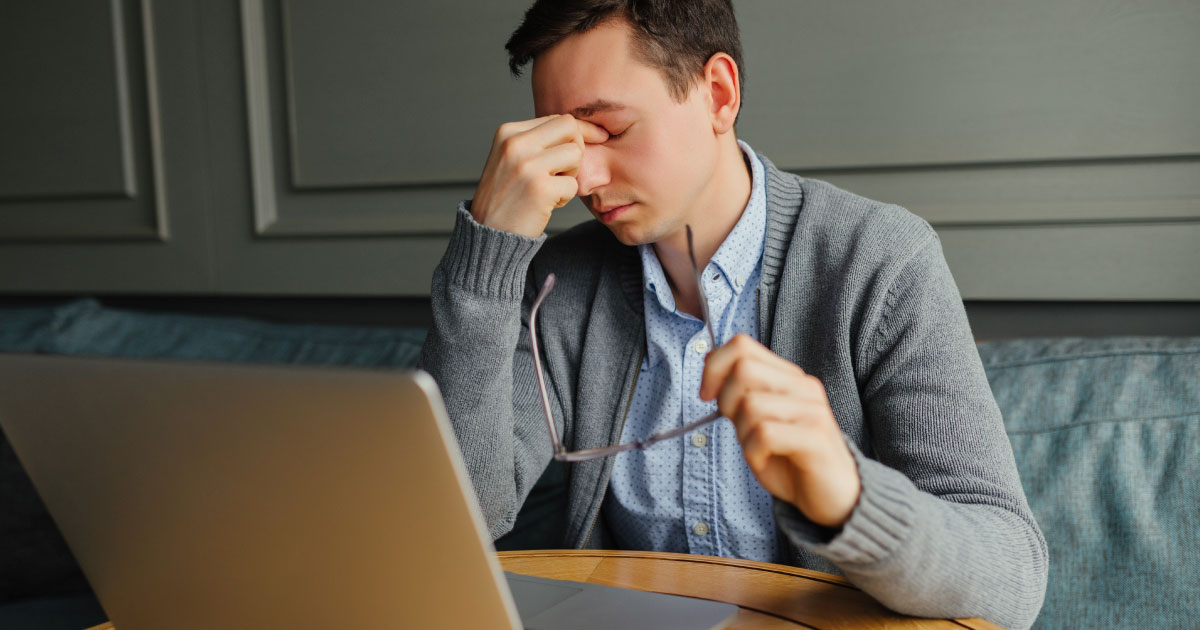 Foto de um homem em sua mesa de trabalho, com um computador à frente, uma das mãos encosta no nariz e os olhos estão fechados. Seu semblante é cansado.