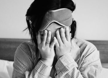 Foto em preto e branco de uma mulher em sua cama, com as mãos na cabeça tapando seus olhos, demonstrando insônia