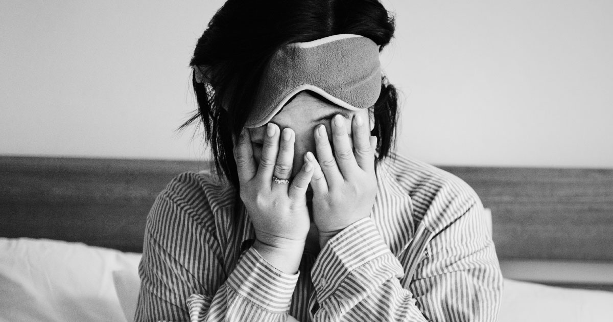 Foto em preto e branco de uma mulher em sua cama, com as mãos na cabeça tapando seus olhos, demonstrando insônia