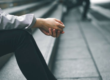 Foto de um homem sentado na rua, só aparecem tronco e pernas, as mãos estão a frente unidas.