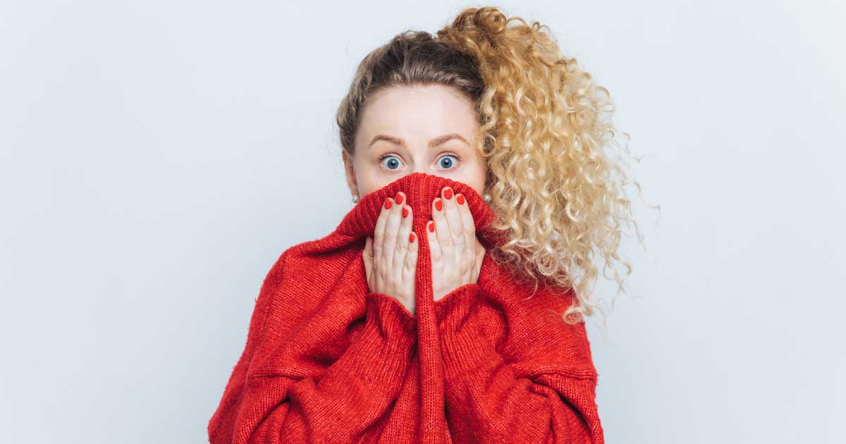 foto de uma mulher loira, com blusa de moletom vermelha, usando a blusa para tapar sua boca. Seus olhos demonstram espanto e medo.