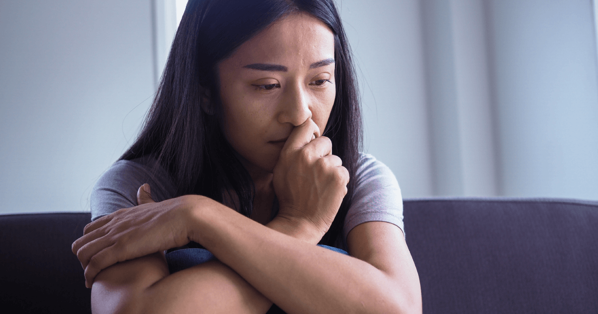 mulher oriental sentada em um sofá com uma das mãos no queixo e semblante ansioso