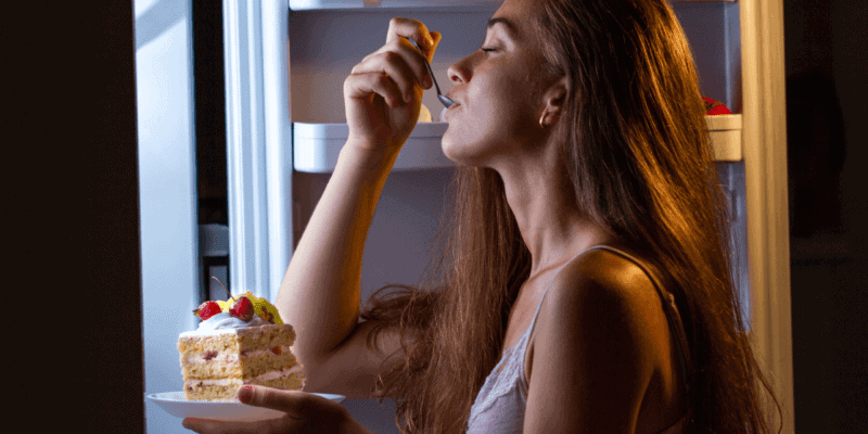 Foto de uma mulher com a geladeira aberta, á noite, comendo um pedaço de bolo de forma compulsiva