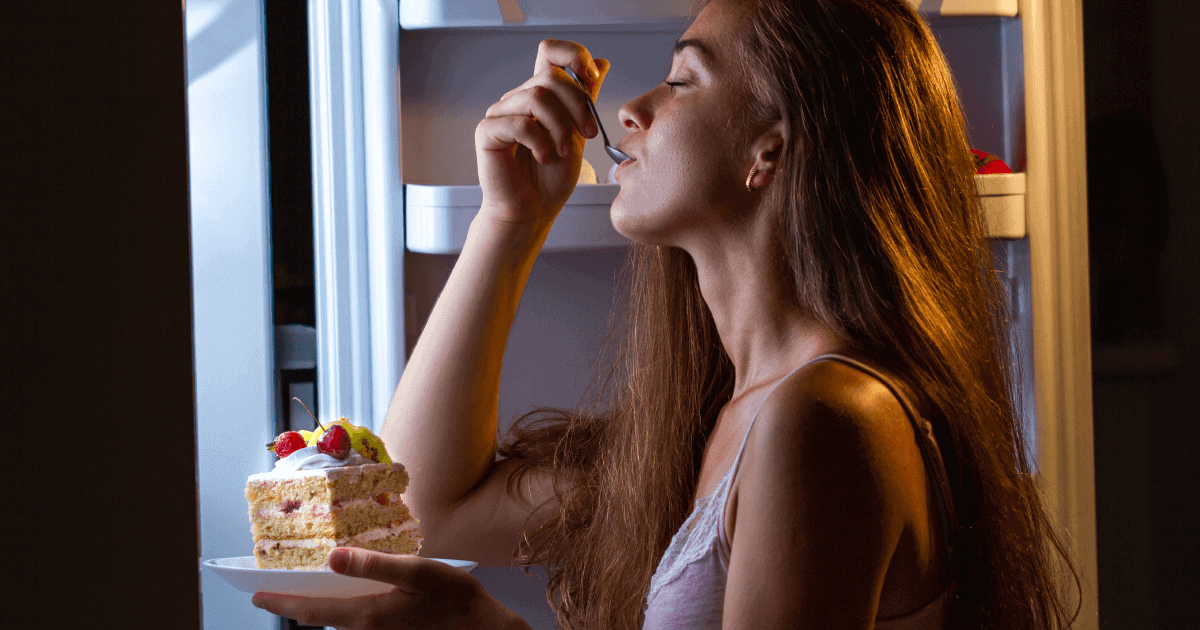 Foto de uma mulher com a geladeira aberta, á noite, comendo um pedaço de bolo de forma compulsiva