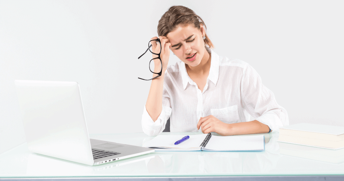 Foto de uma jovem sentada. à sua frete tem um caderno e um computador. Uma das mãos está apoiando a testa e segurando os óculos. o semblante é de dor crônica