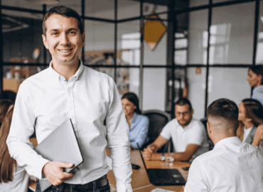 foto de um homem em pé sorrindo e com uma pasta na mão. Atrás dele estão vários colegas de trabalho sentados em uma mesa de reunião