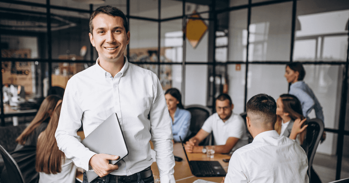 foto de um homem em pé sorrindo e com uma pasta na mão. Atrás dele estão vários colegas de trabalho sentados em uma mesa de reunião