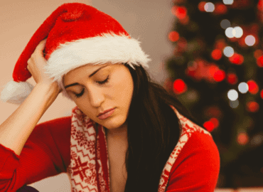 foto de uma mulher sentada na sala, com uma das mãos na cabeça e olhando para baixo com semblante triste. atrás dela tem uma árvore de Natal.