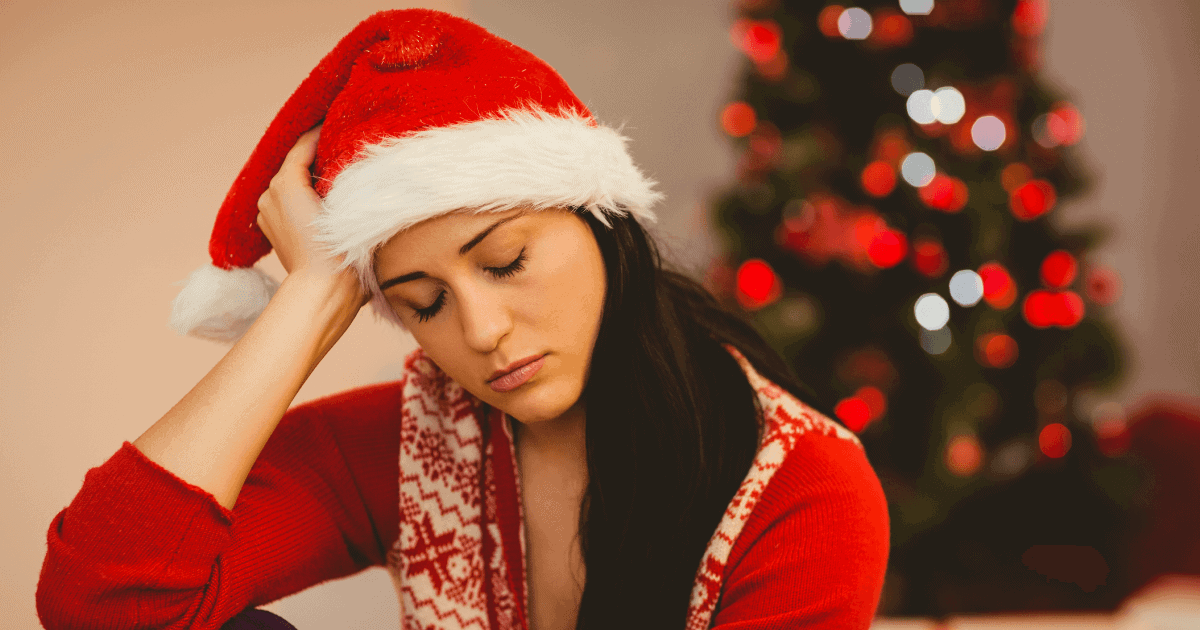 foto de uma mulher sentada na sala, com uma das mãos na cabeça e olhando para baixo com semblante triste. atrás dela tem uma árvore de Natal.